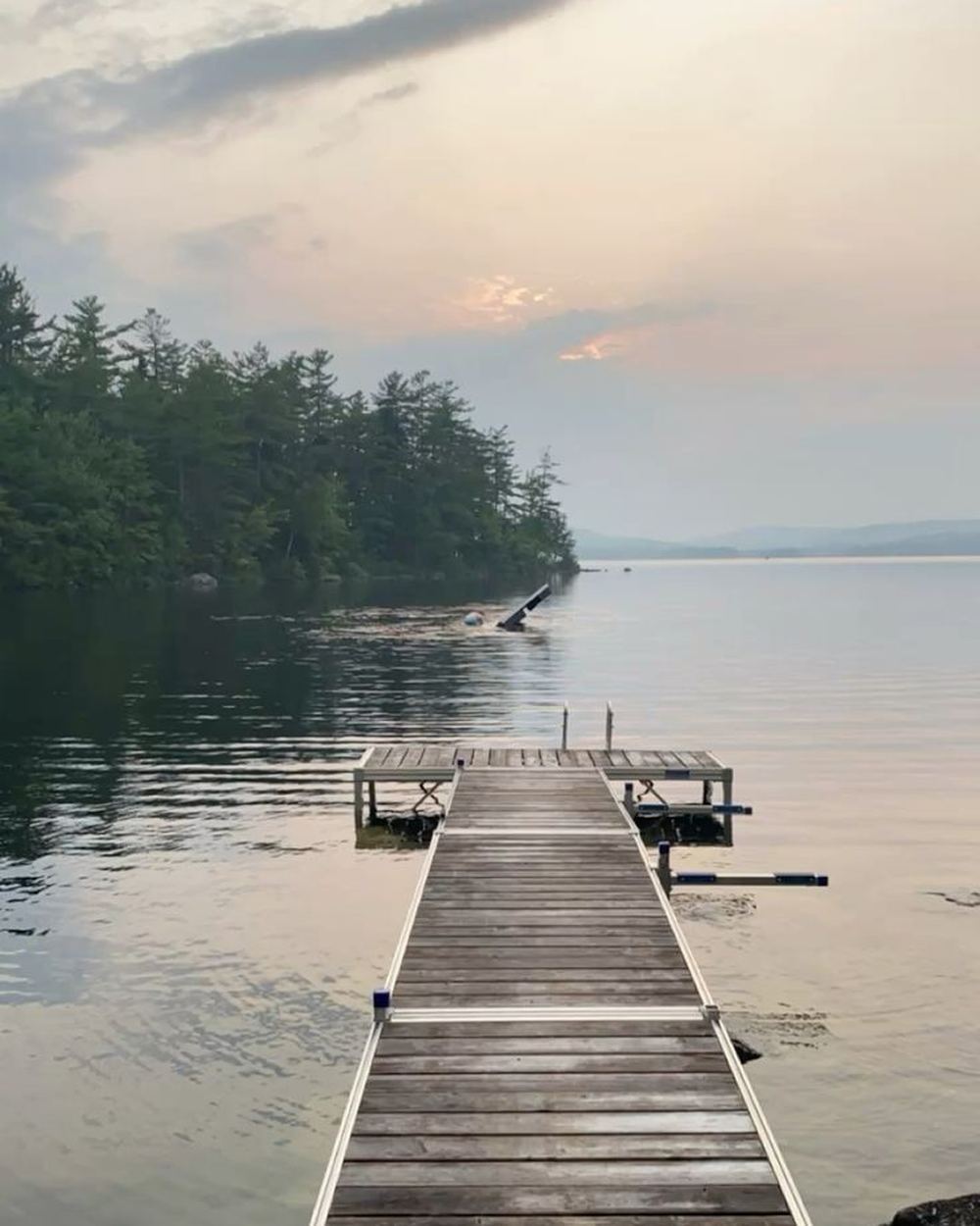 A wooden dock reaching out into a still Maine lake at dusk, pine trees along the far shore